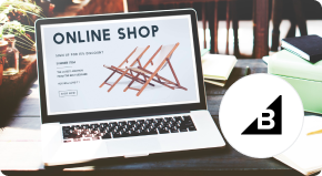 Laptop displaying an “ONLINE SHOP” webpage with crossed-out shopping icons, on a desk beside a round black-and-white B logo; digital commerce.Laptop displaying an “ONLINE SHOP” webpage with crossed-out shopping icons, on a desk beside a round black-and-white B logo; digital commerce.