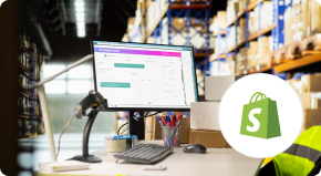 Desktop computer with order management software on screen, barcode scanner, and office supplies in a warehouse; Shopify logo displayed in the foreground.